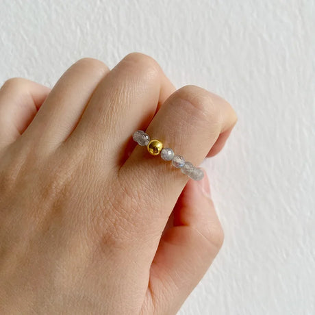 Hand wearing a ring with gold and clear beads on a plain background