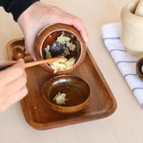 Person using a wooden spoon to mix ingredients in a small wooden bowl on a wooden tray.