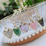 Heart-shaped pendants on gold chains displayed on a decorative stand with a blurred greenery background.