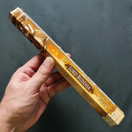 Hand holding a gold 'Lord Buddha' incense stick box against a dark background