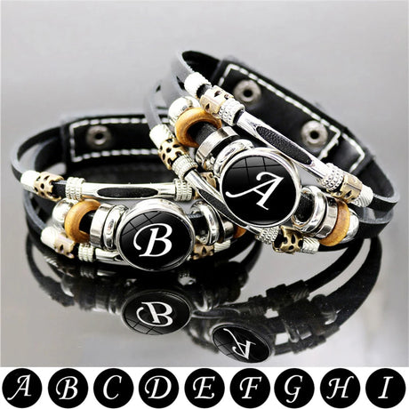 Black leather bracelet with silver and gold charms featuring initials on a gray background.