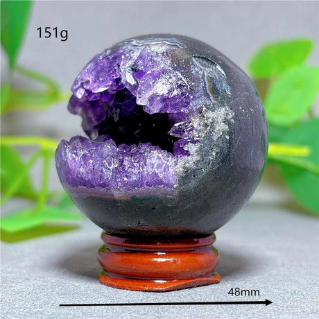 Geode crystal sphere with purple interior on a stand, surrounded by green leaves.