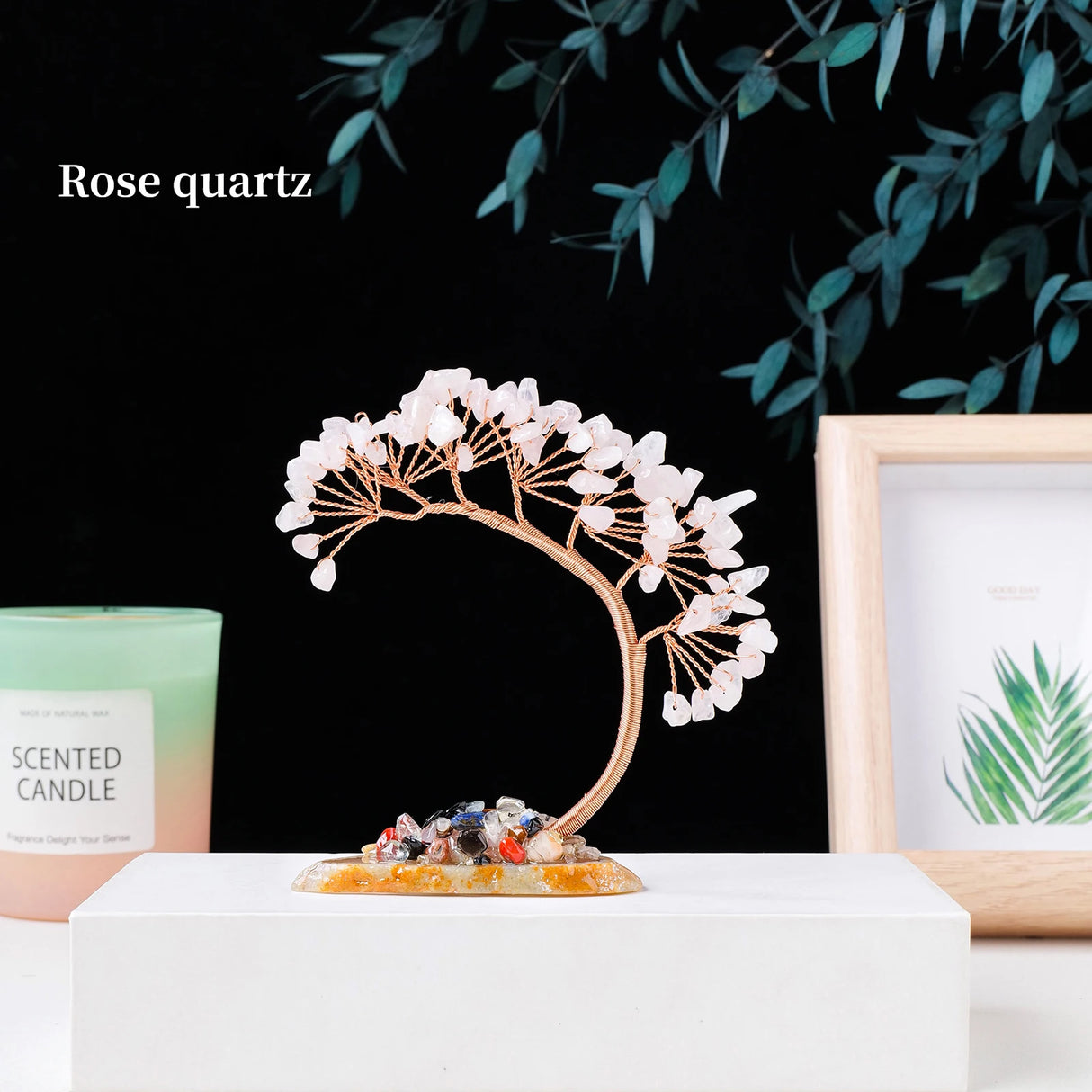 Decorative crystal tree with rose quartz on a white surface, candle, and framed picture in the background.