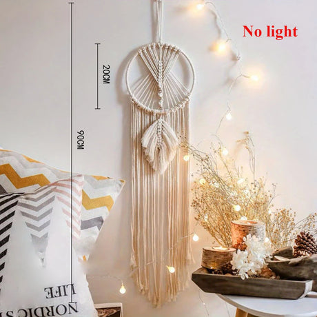 Macrame wall hanging with lights on a white wall, next to decorative pillows and a table with plants.
