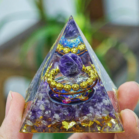 Pyramid-shaped crystal with purple sphere and gold accents held by a hand against a blurred natural background