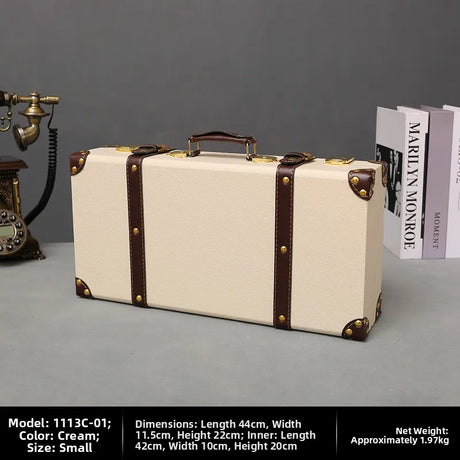 Vintage-style cream suitcase with brown straps on a gray surface, with books and a phone in the background.