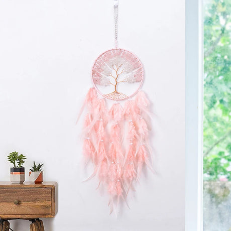 Pink dreamcatcher with feathers hanging on a white wall next to a window with greenery outside.