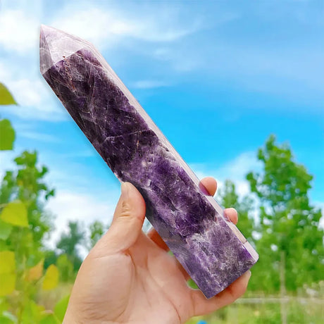 Hand holding a purple crystal against a natural background with trees and blue sky.