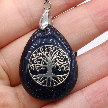 Black teardrop-shaped pendant with silver 'Tree of Life' design held in a hand.