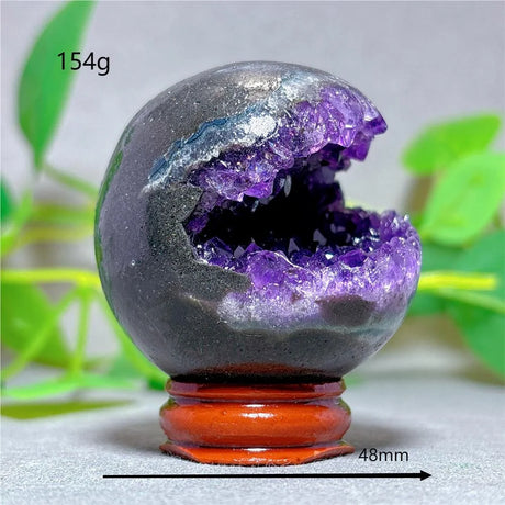 Geode crystal sphere with purple interior on a stand, surrounded by green leaves.