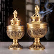 Two ornate gold incense burners with intricate designs on a reflective surface.