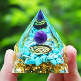 Pyramid-shaped crystal object with a purple ball and turquoise stones held by a hand against a green blurred background