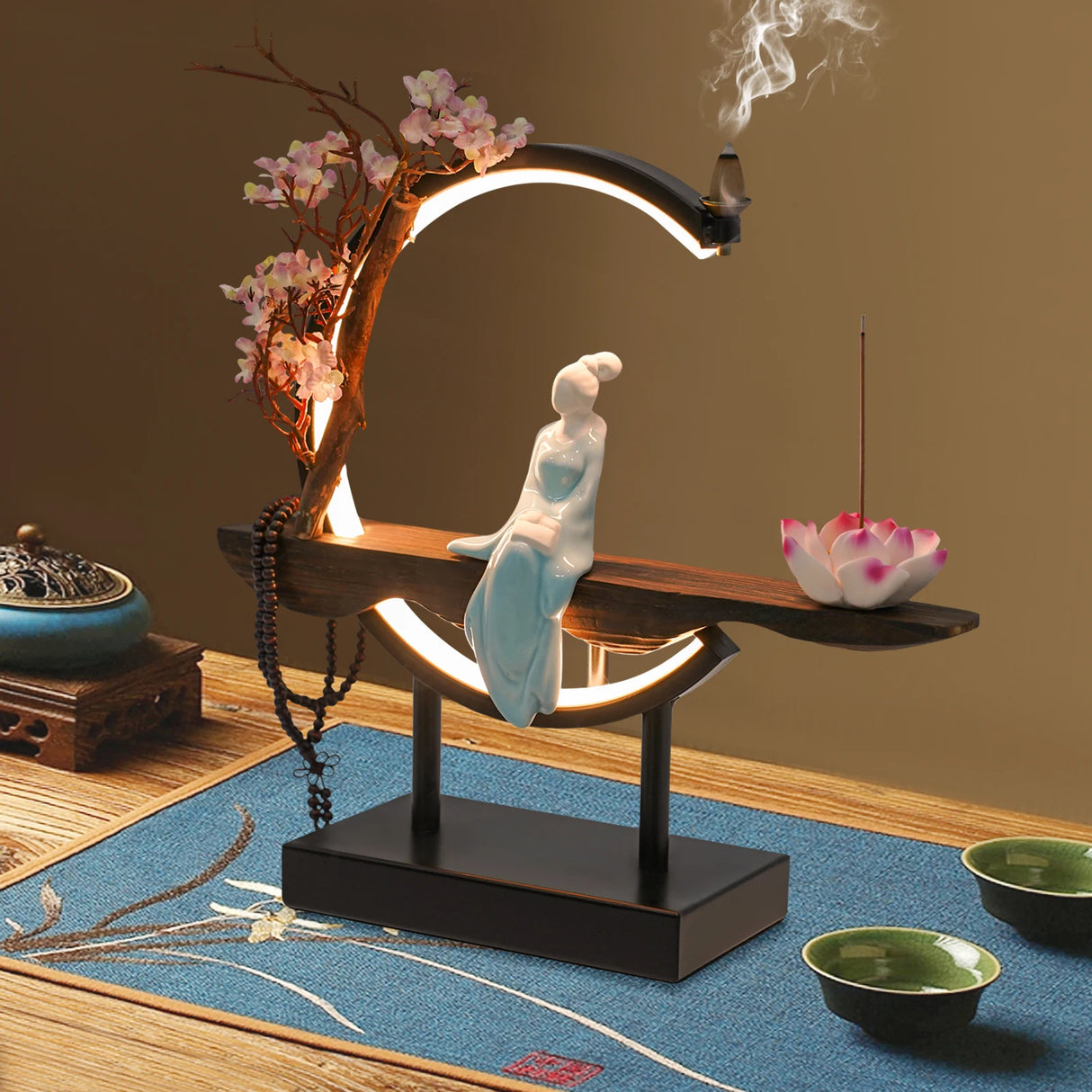 Decorative lamp with a figurine on a wooden platform, surrounded by incense and flowers.