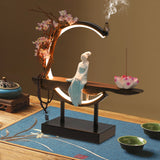 Decorative lamp with a figurine on a wooden platform, surrounded by incense and flowers.