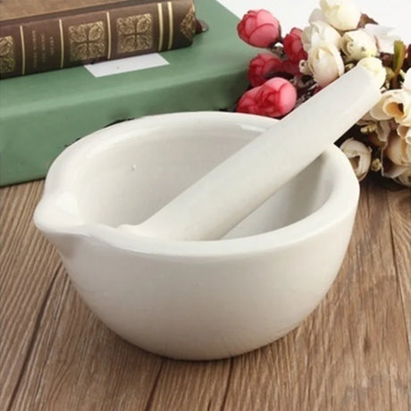 White ceramic mortar and pestle on a wooden surface with flowers and books in the background