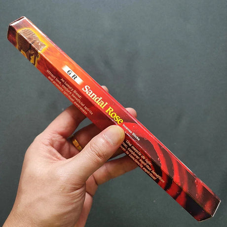 Hand holding a red box of Sandal Rose incense sticks against a dark background