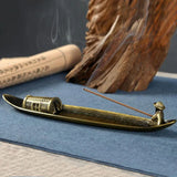 Brass incense burner with incense stick on a blue surface, with wooden elements in the background.