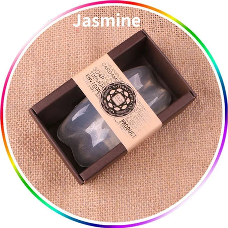 Candle in a box with a label on a textured surface, labeled 'Jasmine'.