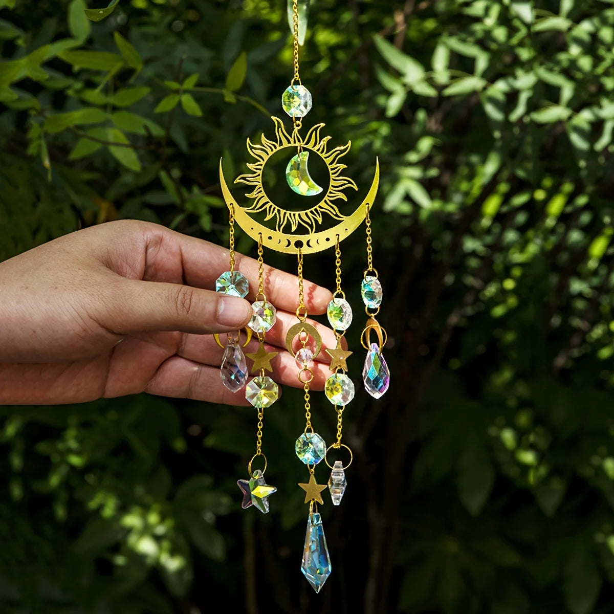 Hand holding a decorative sun and moon ornament with crystals against a green leafy background