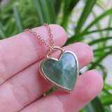 Heart-shaped green stone pendant on a gold chain held in a hand with a blurred green plant background