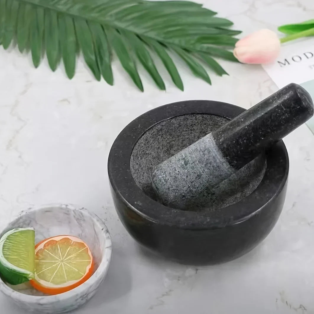 Black stone mortar and pestle on a light surface with lime and orange slices, and green leaves in the background.