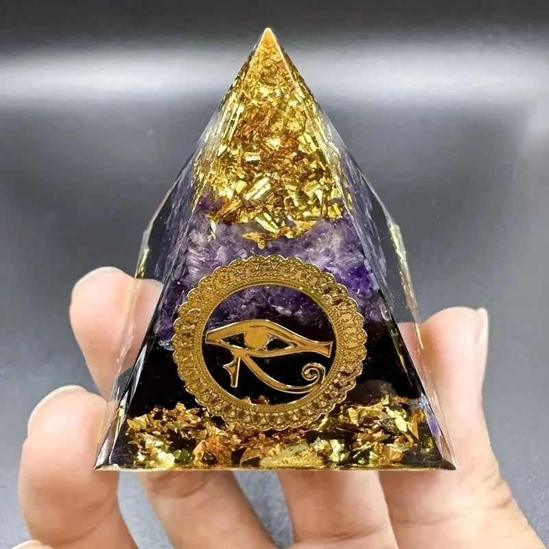 Pyramid-shaped crystal with gold and purple colors, featuring an eye symbol, held by a hand against a gray background.