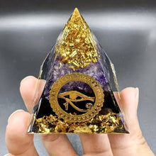 Pyramid-shaped crystal with gold and purple colors, featuring an eye symbol, held by a hand against a gray background.
