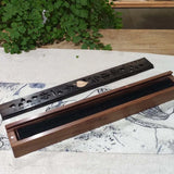 Wooden incense burner with engraved design on a textured surface with greenery in the background