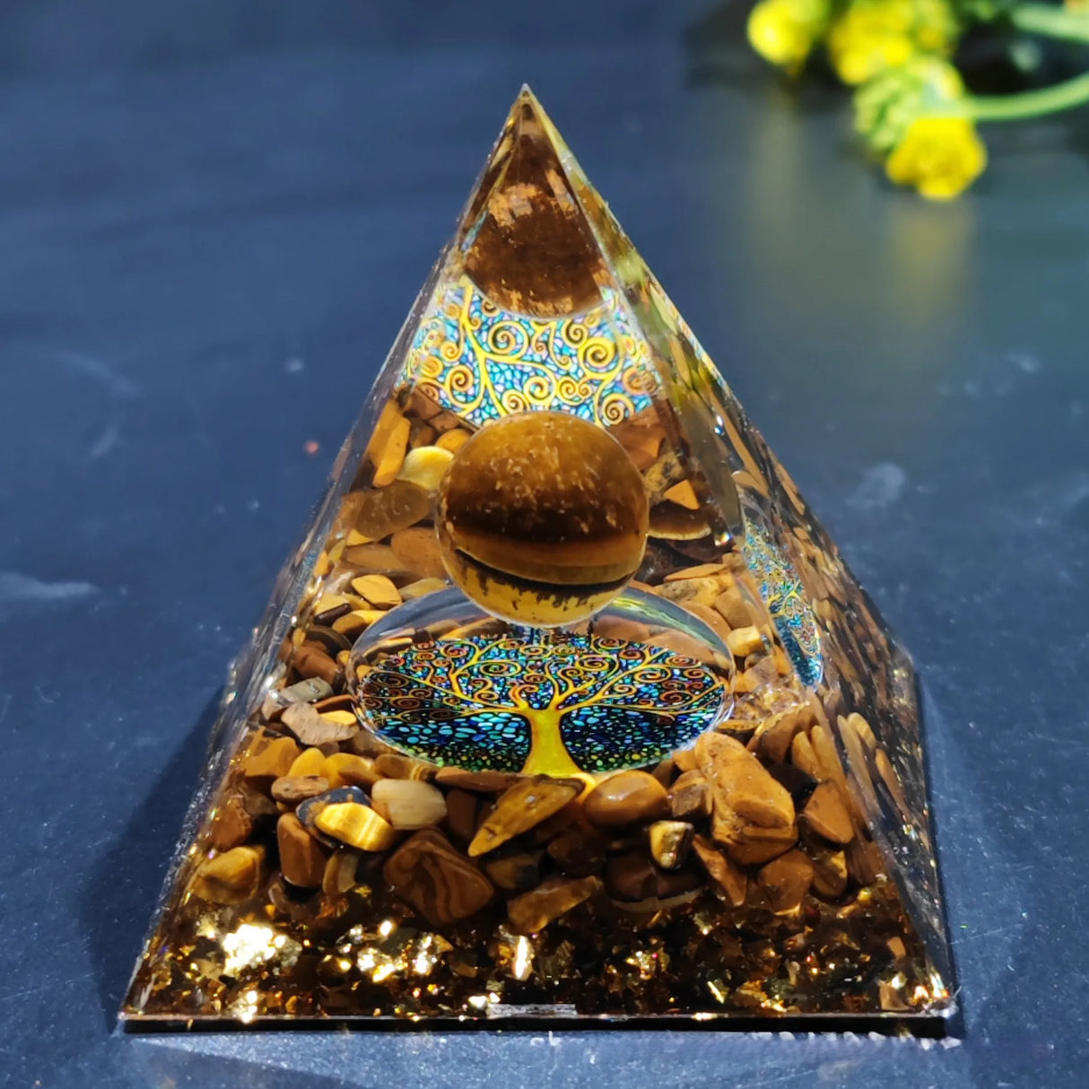 Pyramid-shaped decorative item with stones and a tree of life design on a dark surface.