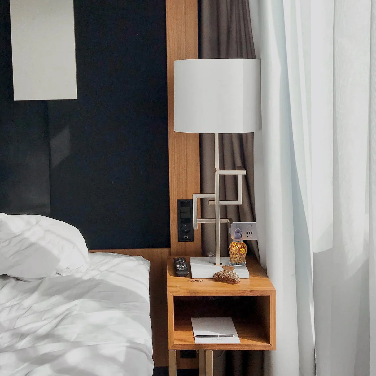 Wooden nightstand with lamp and remote control next to a bed in a room with curtains.