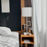 Wooden nightstand with lamp and remote control next to a bed in a room with curtains.