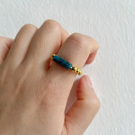 Hand wearing a ring with blue beads and gold accents on a light background