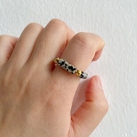 Hand wearing a ring with black, white, and gold beads on a plain background