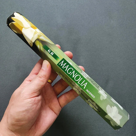 Hand holding a green incense stick packaging with magnolia design against a gray background
