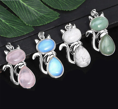 Four cat-shaped pendants with colorful stones on a dark surface with green leaves.