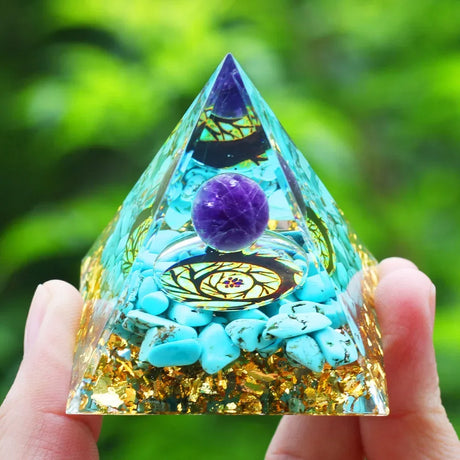 Pyramid-shaped crystal with purple sphere and stones held by a hand against a green blurred background