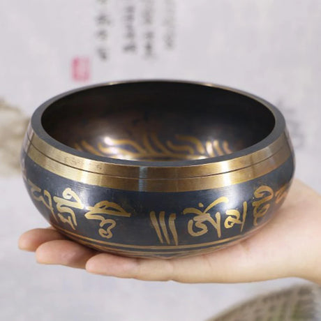 Hand holding a black singing bowl with gold text against a blurred background