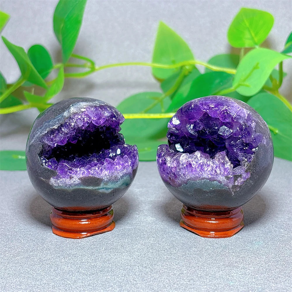 Two purple crystal balls with orange bases on a light gray surface with green leaves in the background.