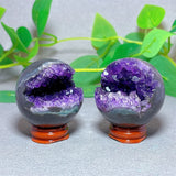 Two purple crystal balls with orange bases on a light gray surface with green leaves in the background.