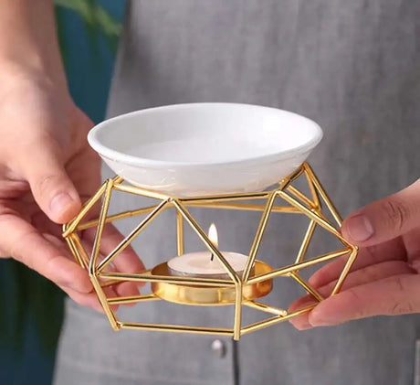 Gold geometric candle holder with a lit candle inside, held by two hands.