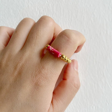 Hand wearing a pink beaded ring with gold beads on a white background