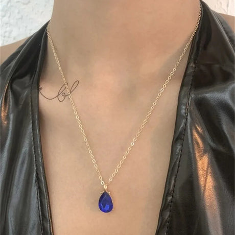 Gold necklace with a blue teardrop pendant worn by a person in a black leather jacket.