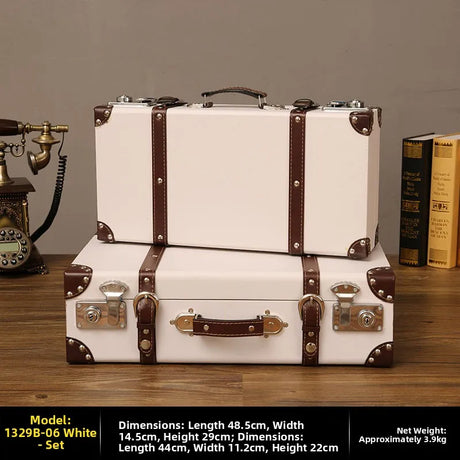 Two vintage-style white suitcases with brown straps on a wooden surface, with a phone and books in the background.