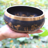 Hand holding a black singing bowl with gold inscriptions against a blurred natural background