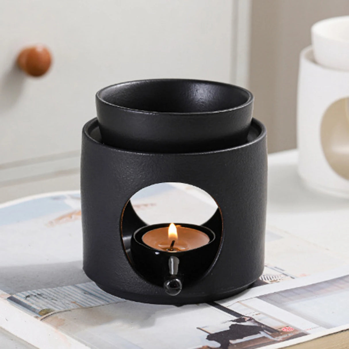Black ceramic oil burner with a lit candle inside on a magazine