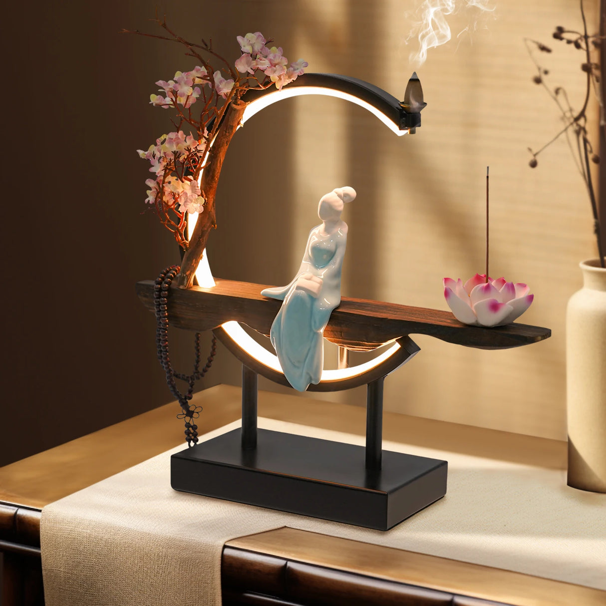 Decorative table lamp with a figurine on a stand, surrounded by flowers and incense.