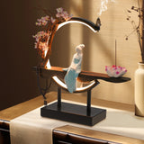 Decorative table lamp with a figurine on a stand, surrounded by flowers and incense.
