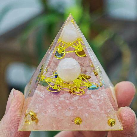 Pyramid-shaped crystal with pink base and gold accents held by a hand against a blurred natural background