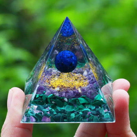 Pyramid-shaped crystal with blue and green elements held in a hand against a blurred green background