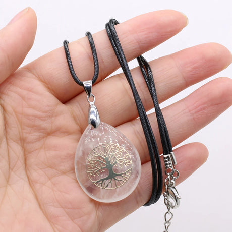 Clear teardrop-shaped pendant with tree of life design on a black cord held in a hand.